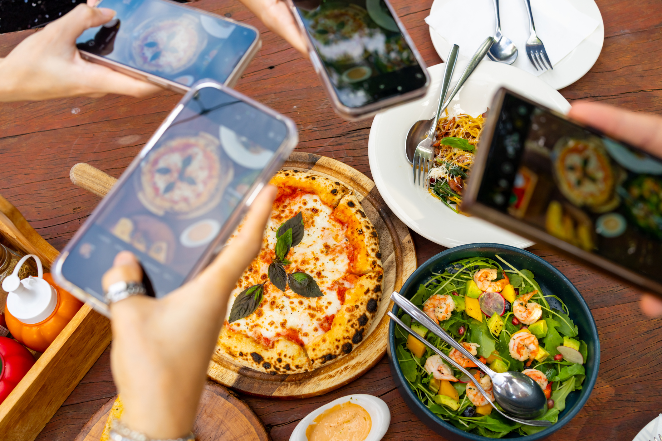 Group of hands holding cell phones and taking photos of food on a table.